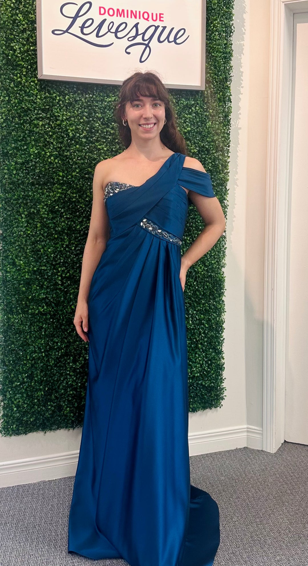 Woman in a blue one-shoulder dress standing in front of a green leafy wall with a sign above.
