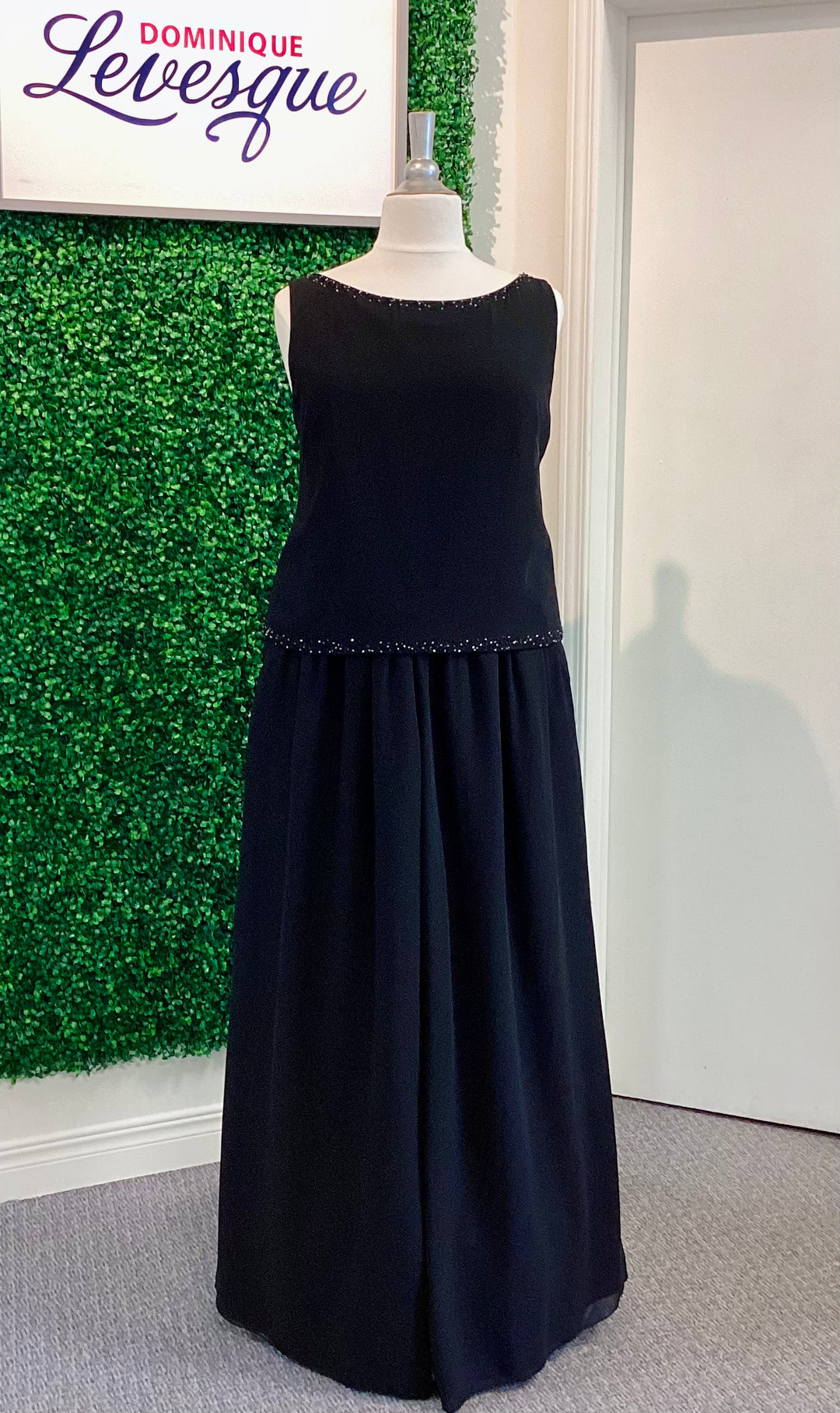 Black dress on a mannequin against a green wall with 'Dominique Levesque' branding.