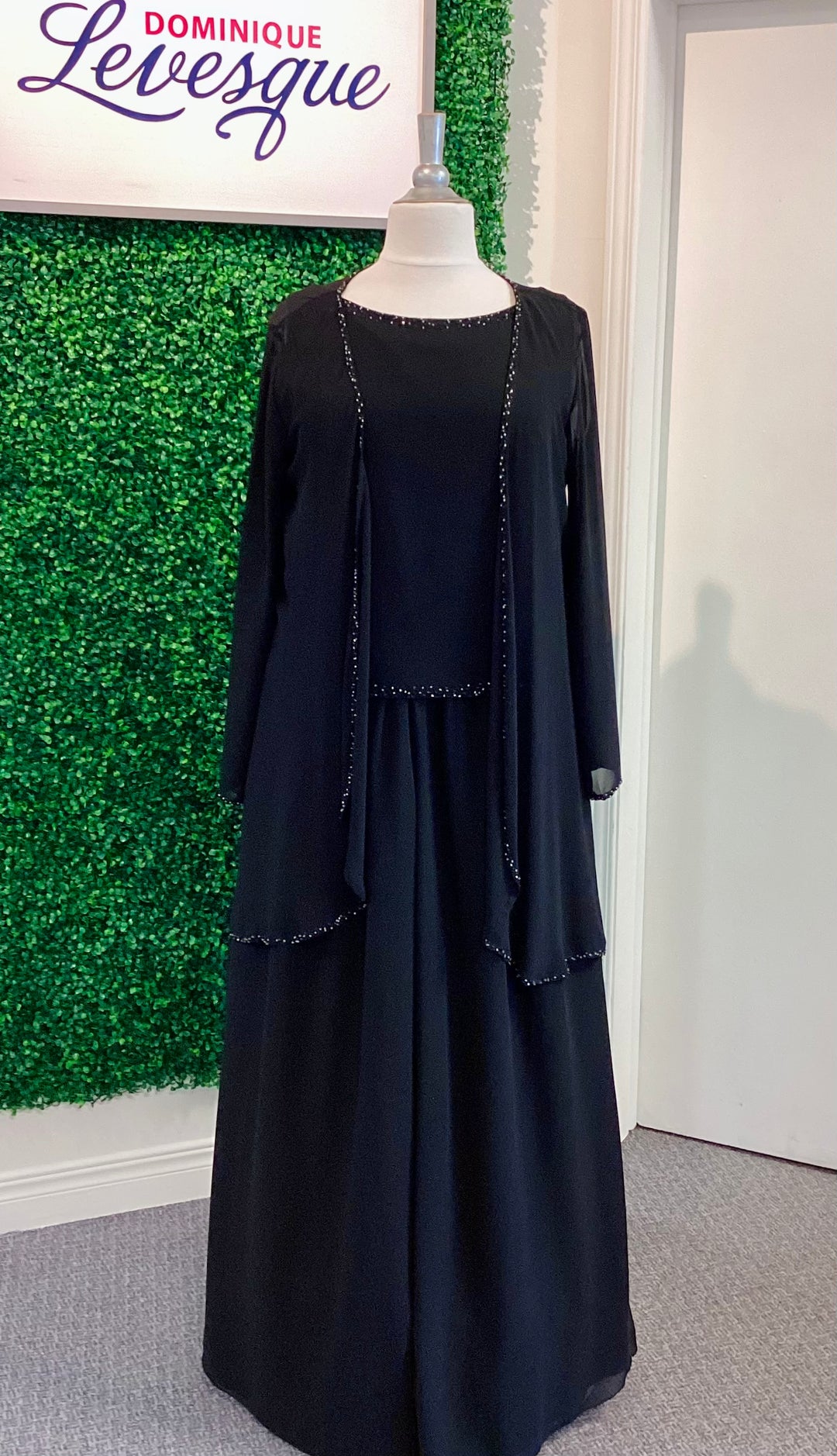 Black pants and top with vest on a mannequin against a green wall with &