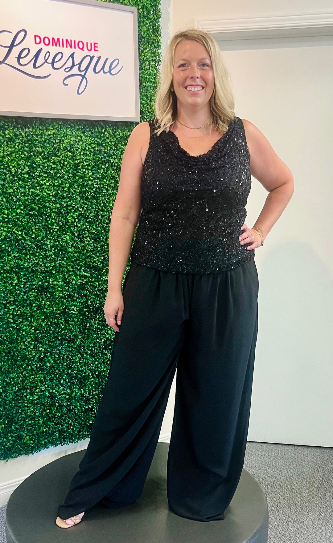 Woman wearing a black sequin top and wide-leg pants standing in front of a green wall with a sign that reads 'Dominique Levesque'.