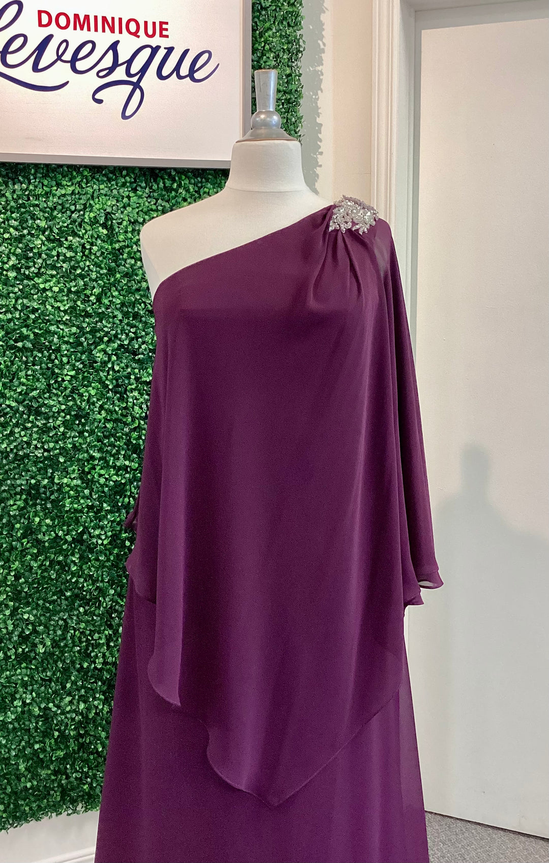 Plum one-shoulder dress on a mannequin against a green leafy background with &