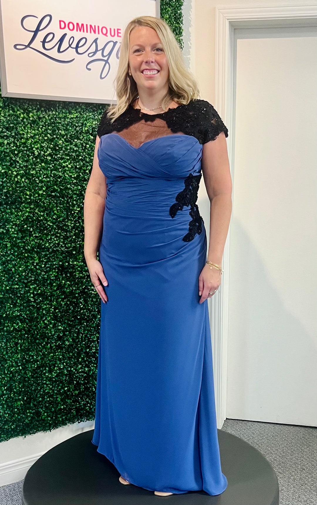 Woman in a blue evening gown with black lace details standing in front of a green wall and white door.