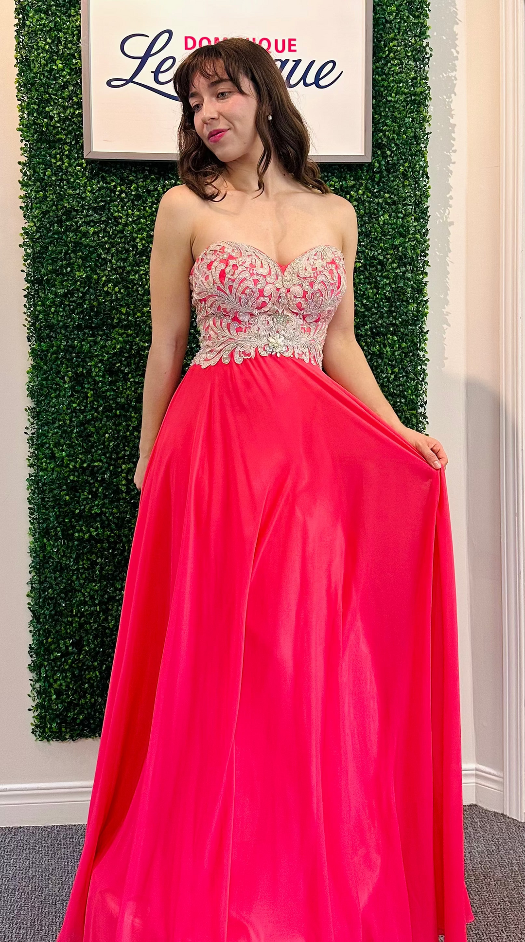 Woman in a pink evening gown with intricate top design against a green leafy background.