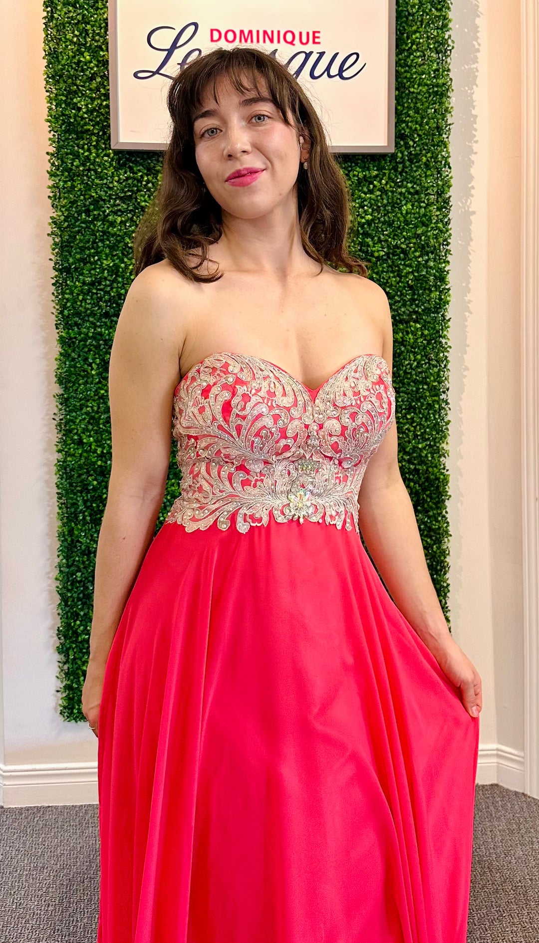 Woman in a pink evening gown with intricate top design against a green leafy background.