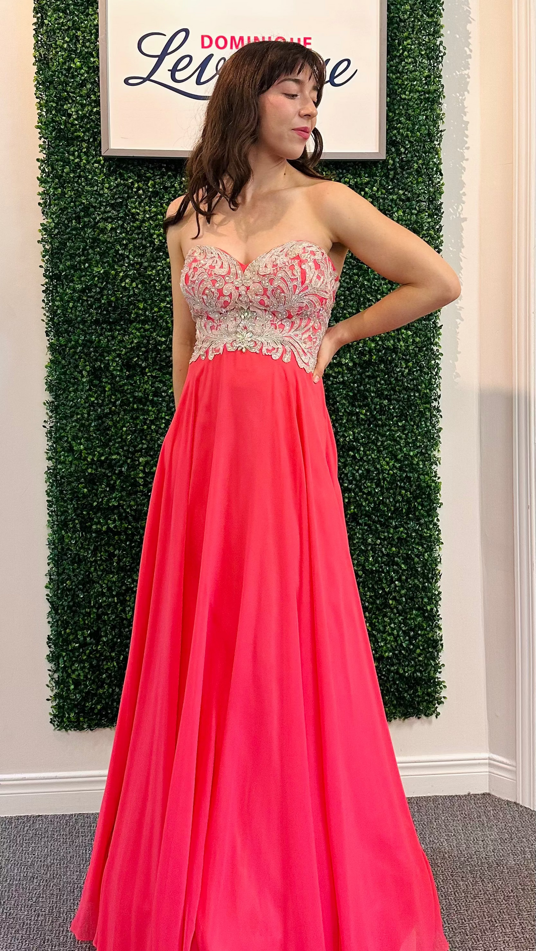 Woman in a pink evening gown with intricate top design against a green leafy background.