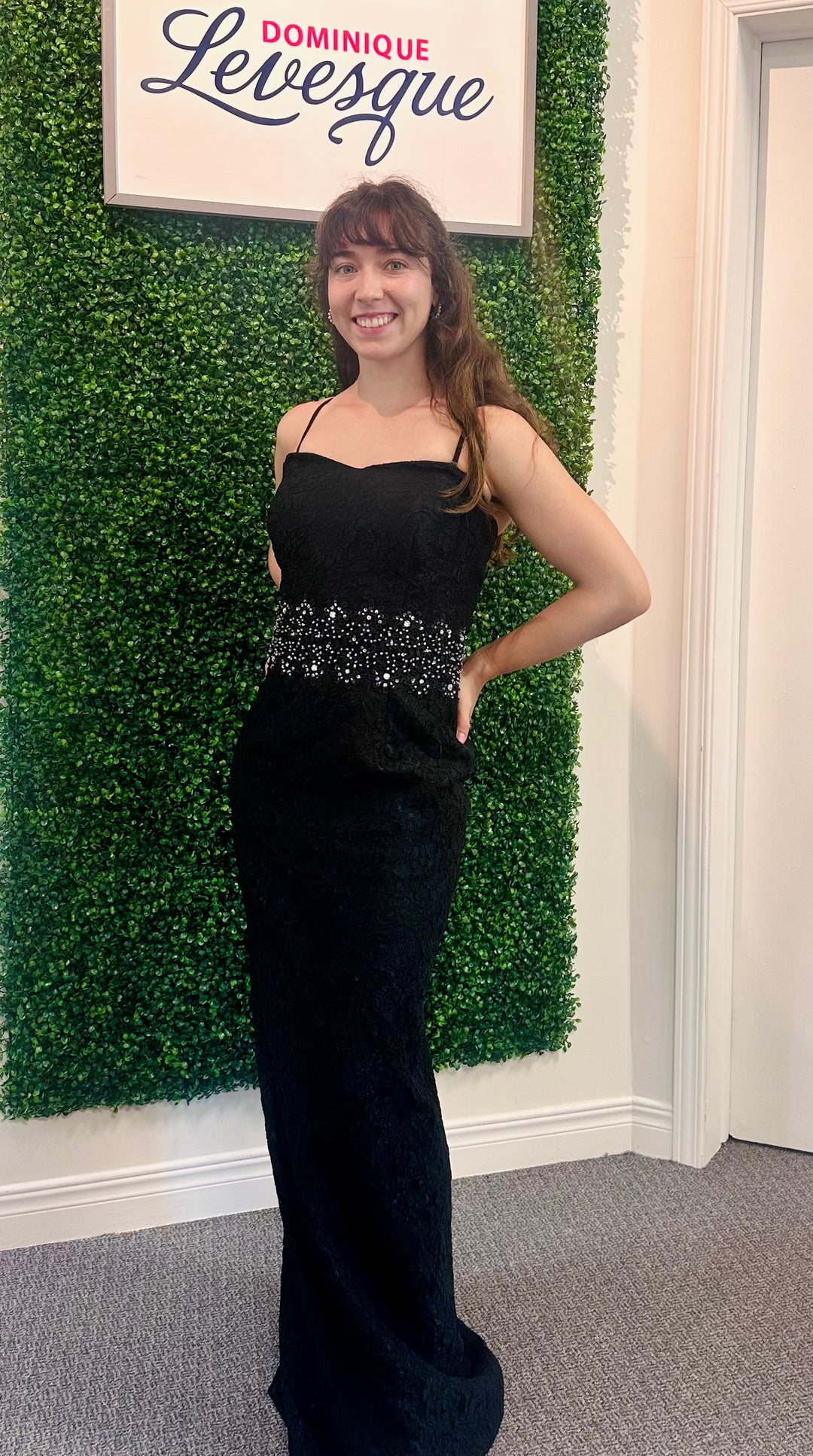 Woman in a black evening gown standing in front of a green leafy wall with a sign above.