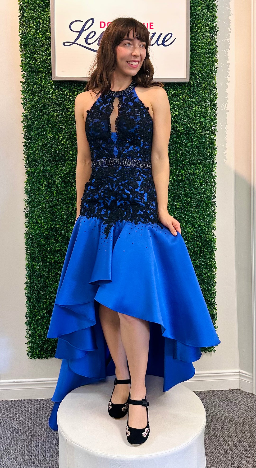 Woman in a blue and black dress standing against a green bush backdrop with a sign above.