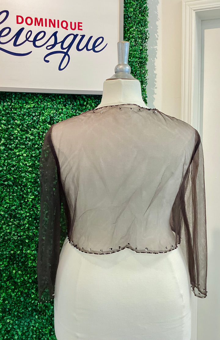 Mannequin wearing a sheer brown top with long sleeves against a green leafy background, featuring the brand &