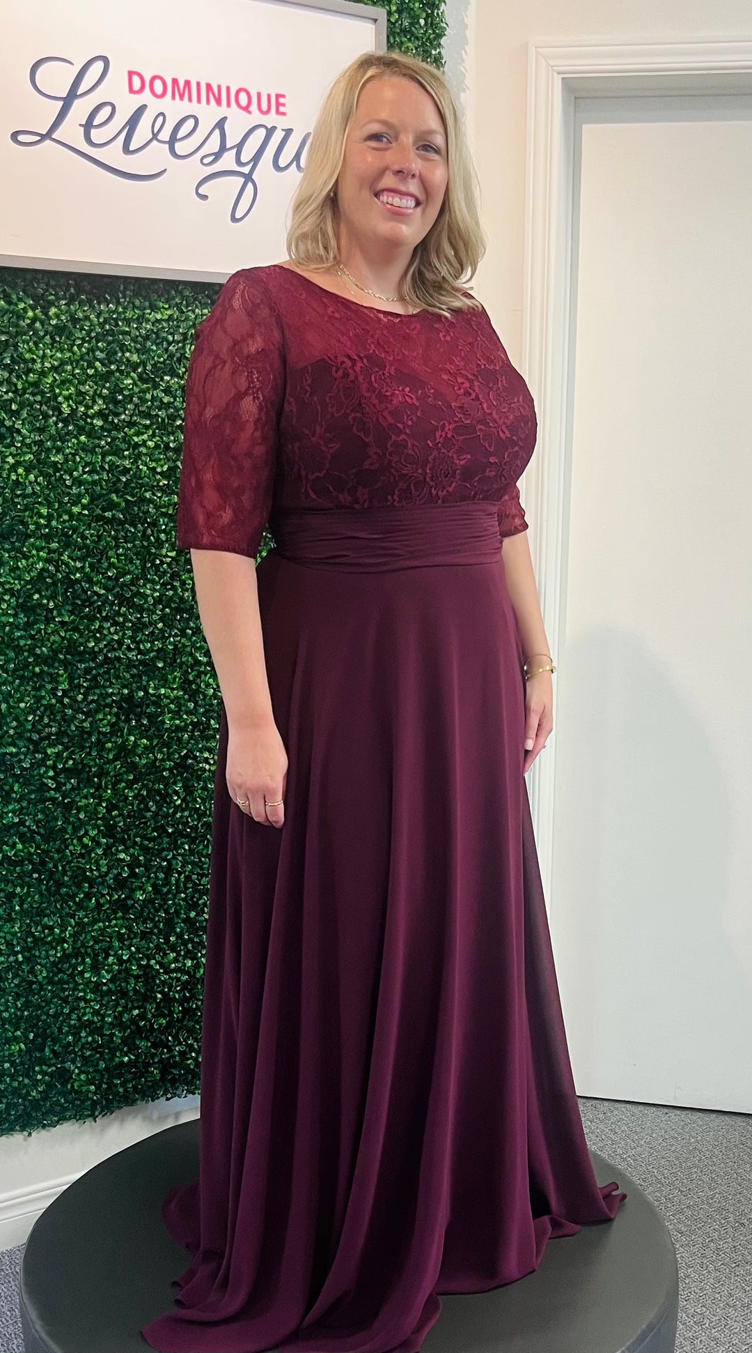 Woman wearing a burgundy evening gown with lace detailing in front of a green wall and white door.