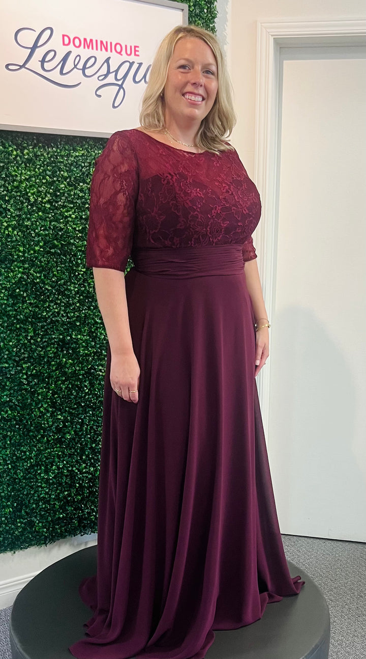 Woman wearing a burgundy evening gown with lace detailing in front of a green wall and white door.