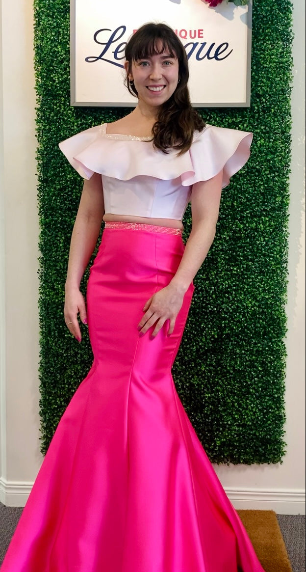 Woman in a baby pink top with ruffled sleeves and a hot pink mermaid-style skirt standing against a green leafy background.