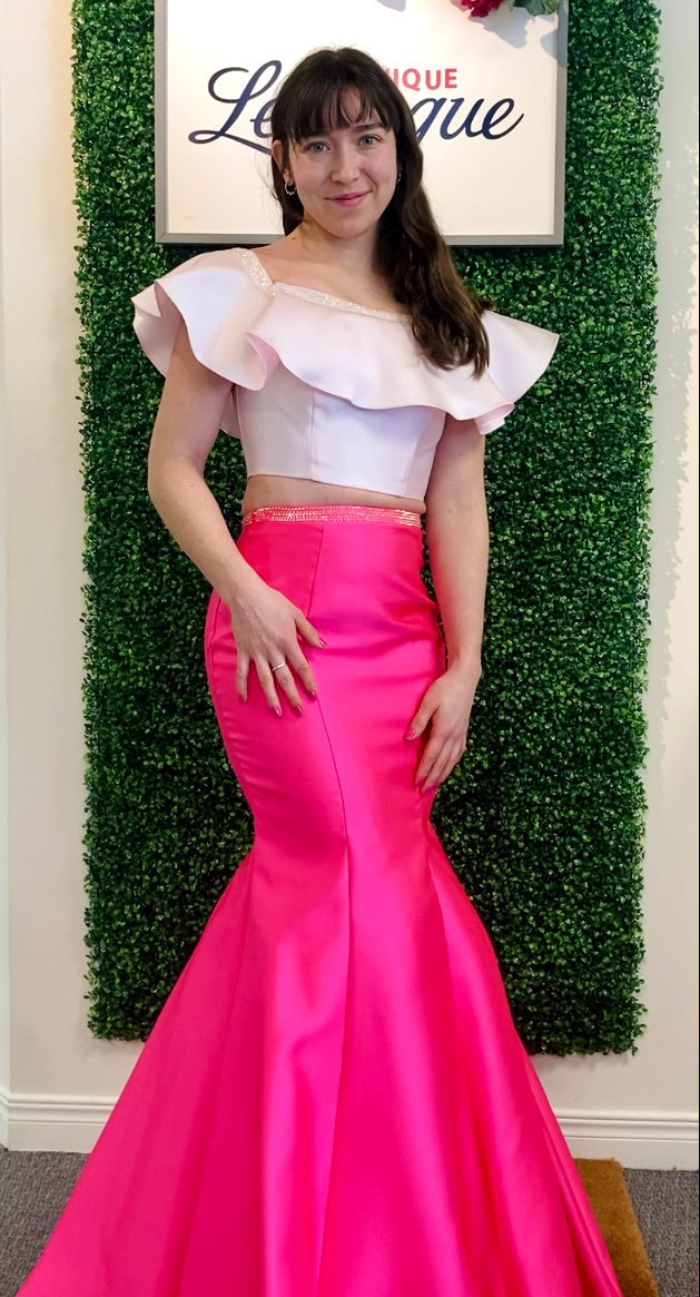 Woman in a baby pink top with ruffled sleeves and a hot pink mermaid-style skirt standing against a green leafy background.