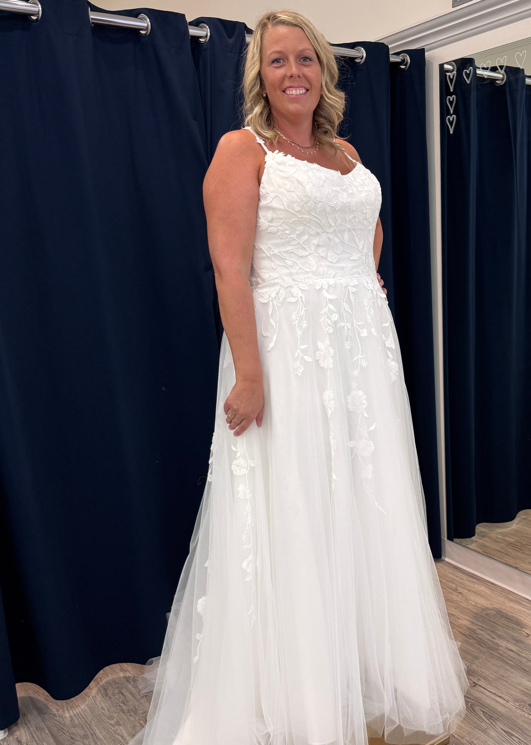 Woman wearing a white floral wedding dress in a fitting room with navy curtains.