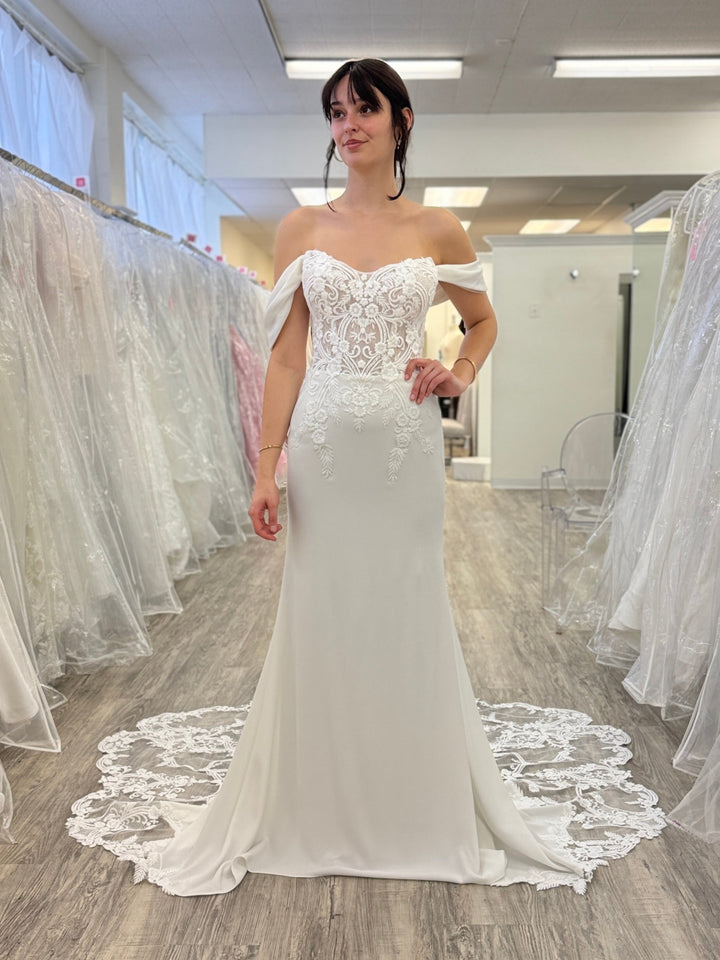 Woman in a white wedding dress standing in a bridal shop with other dresses on racks.