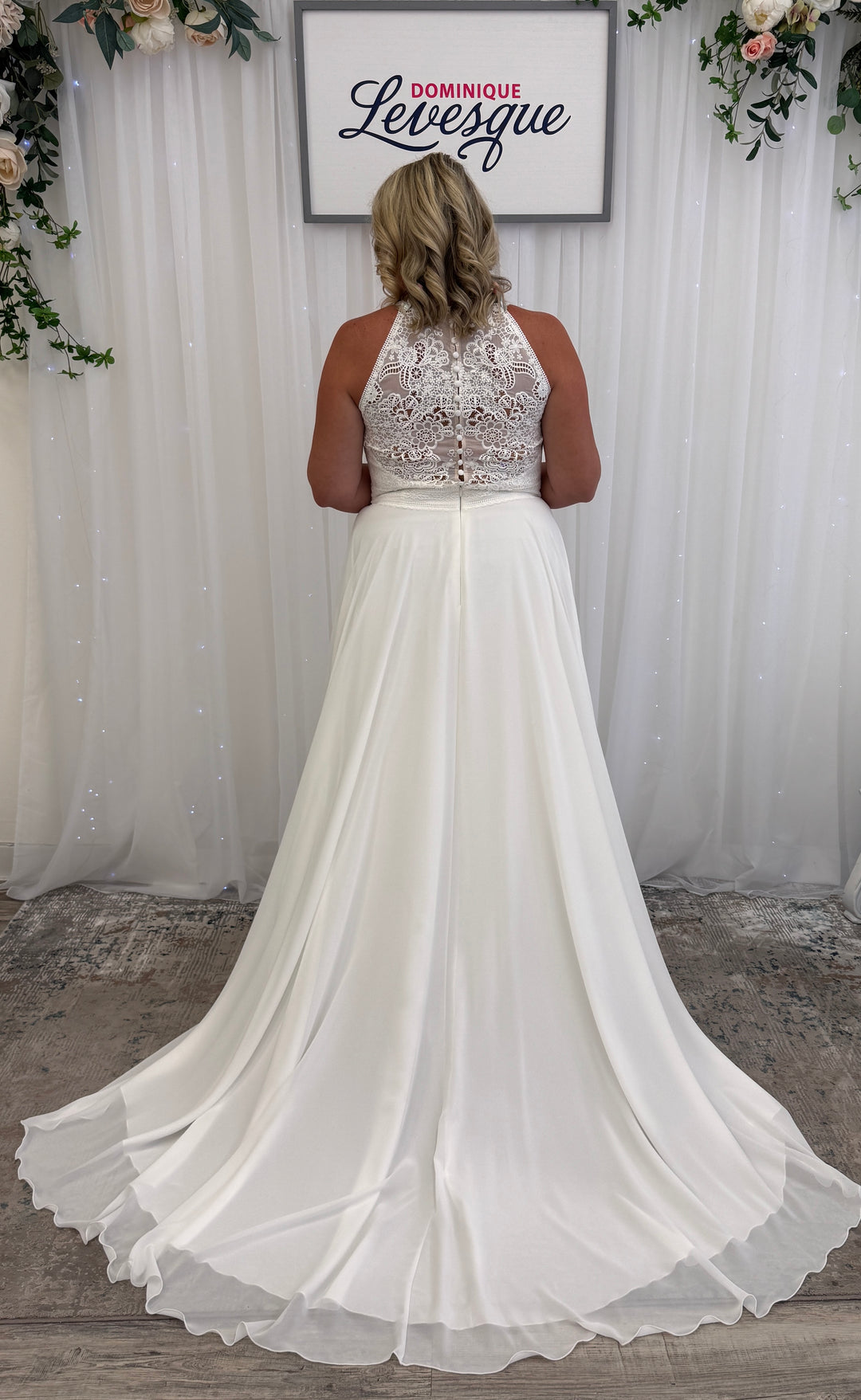 White wedding dress with lace top and flowing skirt in front of a white curtain with floral decorations.