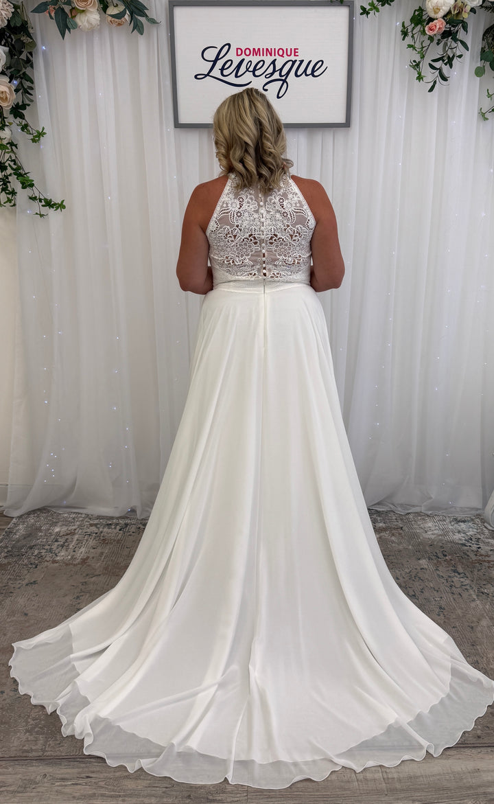 White wedding dress with lace top and flowing skirt in front of a white curtain with floral decorations.