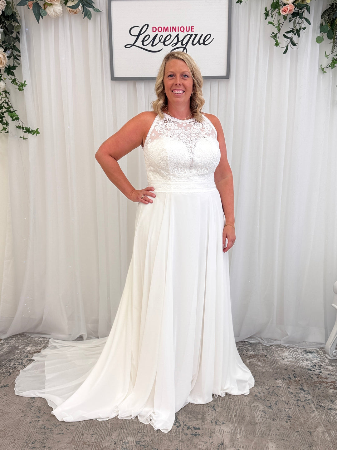 Woman wearing a white plus-size wedding dress in a studio setting with 'Dominique Levesque' branding.