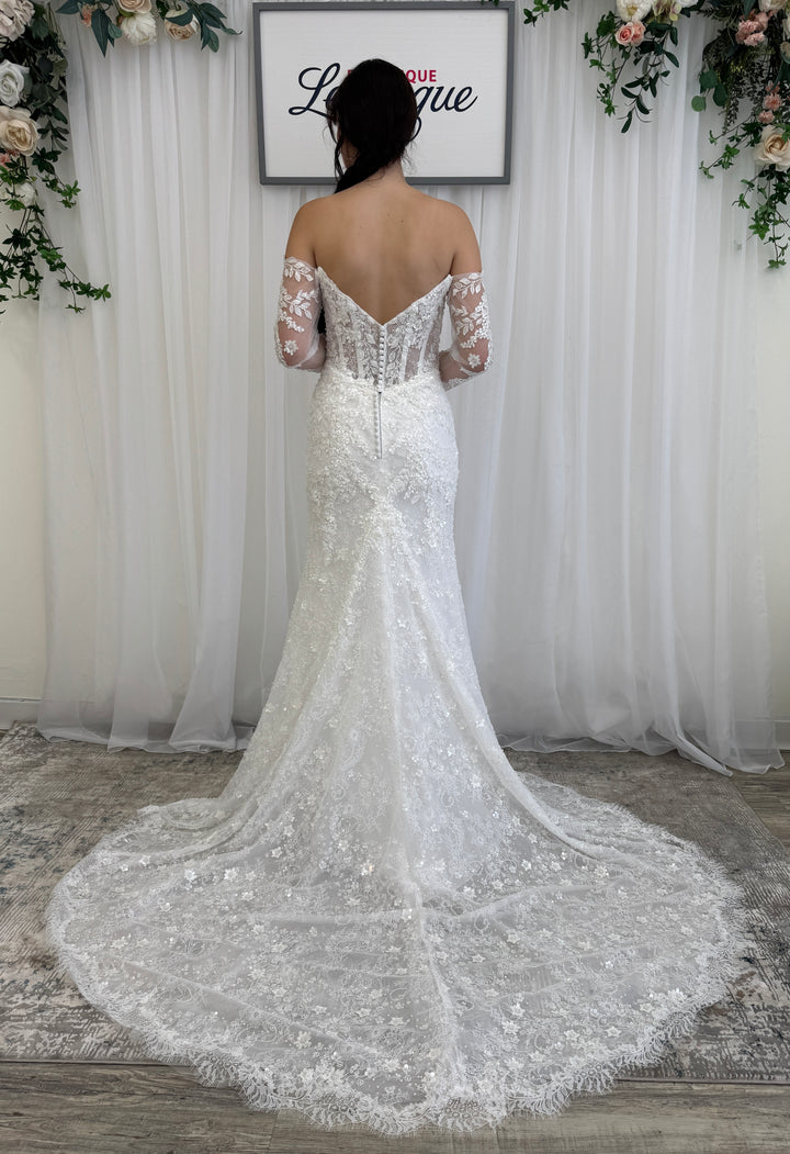 Woman in a white lace wedding dress standing in front of a floral decorated backdrop.