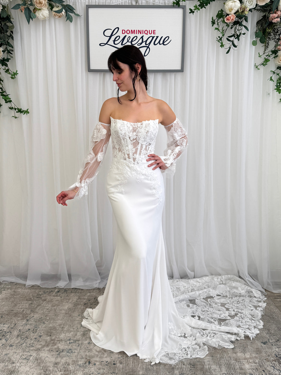 Woman wearing a white wedding dress with lace detailed top and lace sleeves in front of a white curtain with floral decorations.