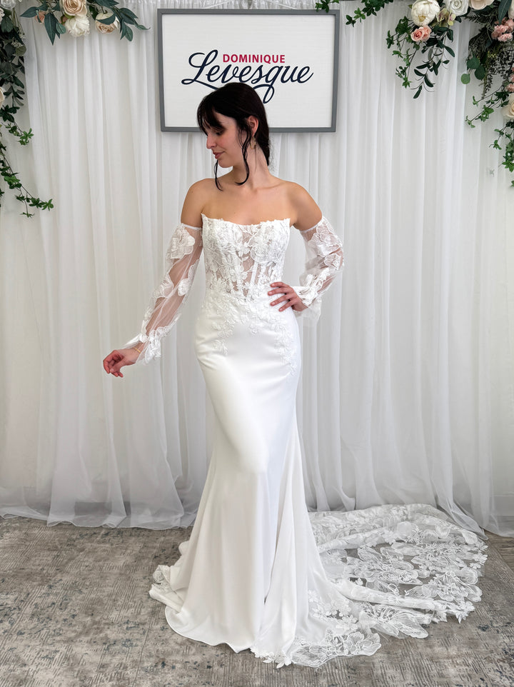 Woman wearing a white wedding dress with lace detailed top and lace sleeves in front of a white curtain with floral decorations.