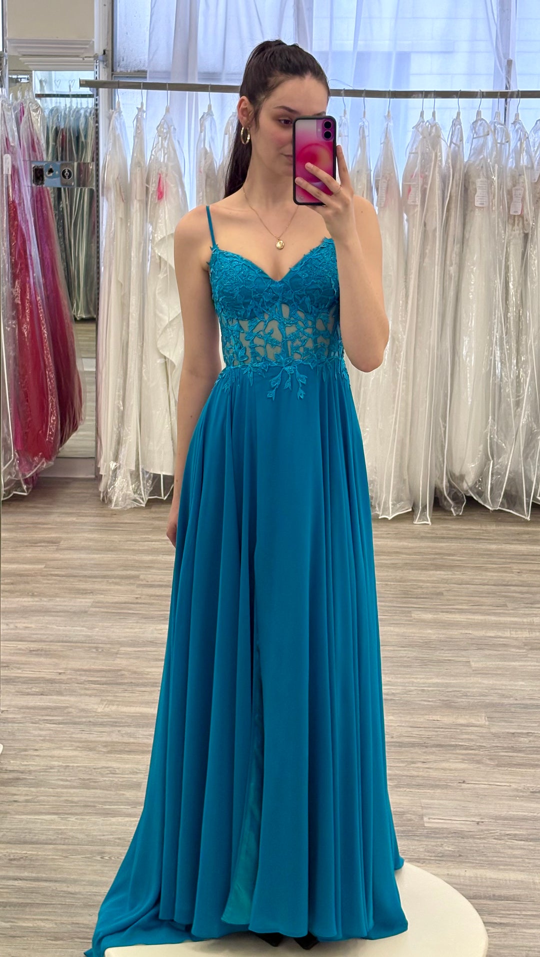 Woman wearing a teal gown in a fitting room with mirrors and dresses.