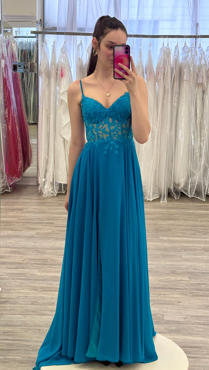 Woman wearing a teal gown in a fitting room with mirrors and dresses.