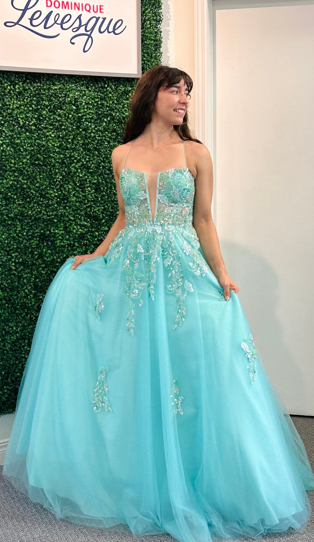 Woman in a light blue evening gown with floral embellishments, standing against a green wall with 'Dominique Levesque' branding.