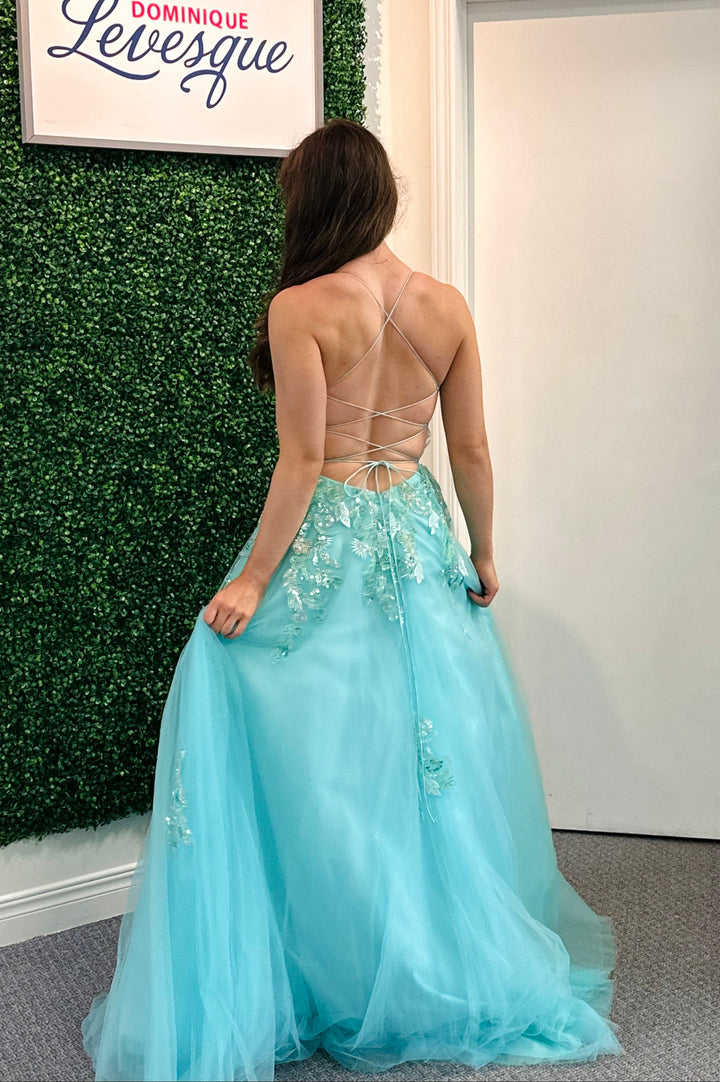 Woman in a light blue evening gown with floral details, standing against a green leafy wall.