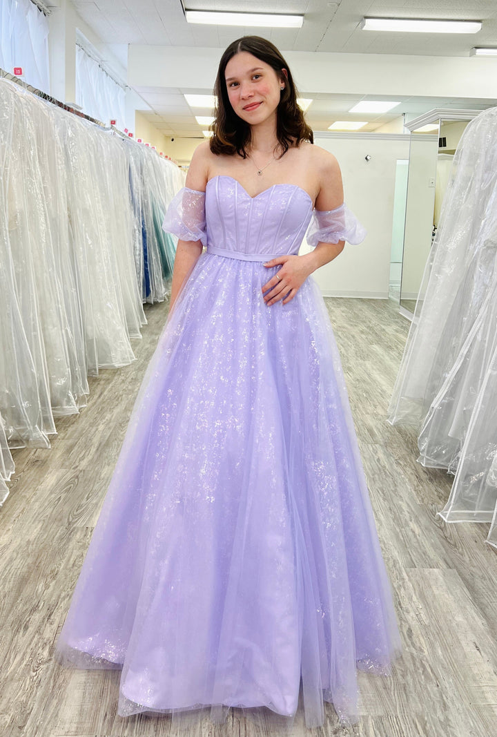 Woman in a lavender evening gown standing in a fitting room with white dresses.