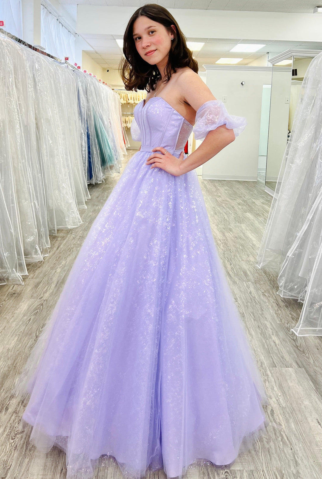 Woman in a lavender prom dress standing in a fitting room with other dresses.