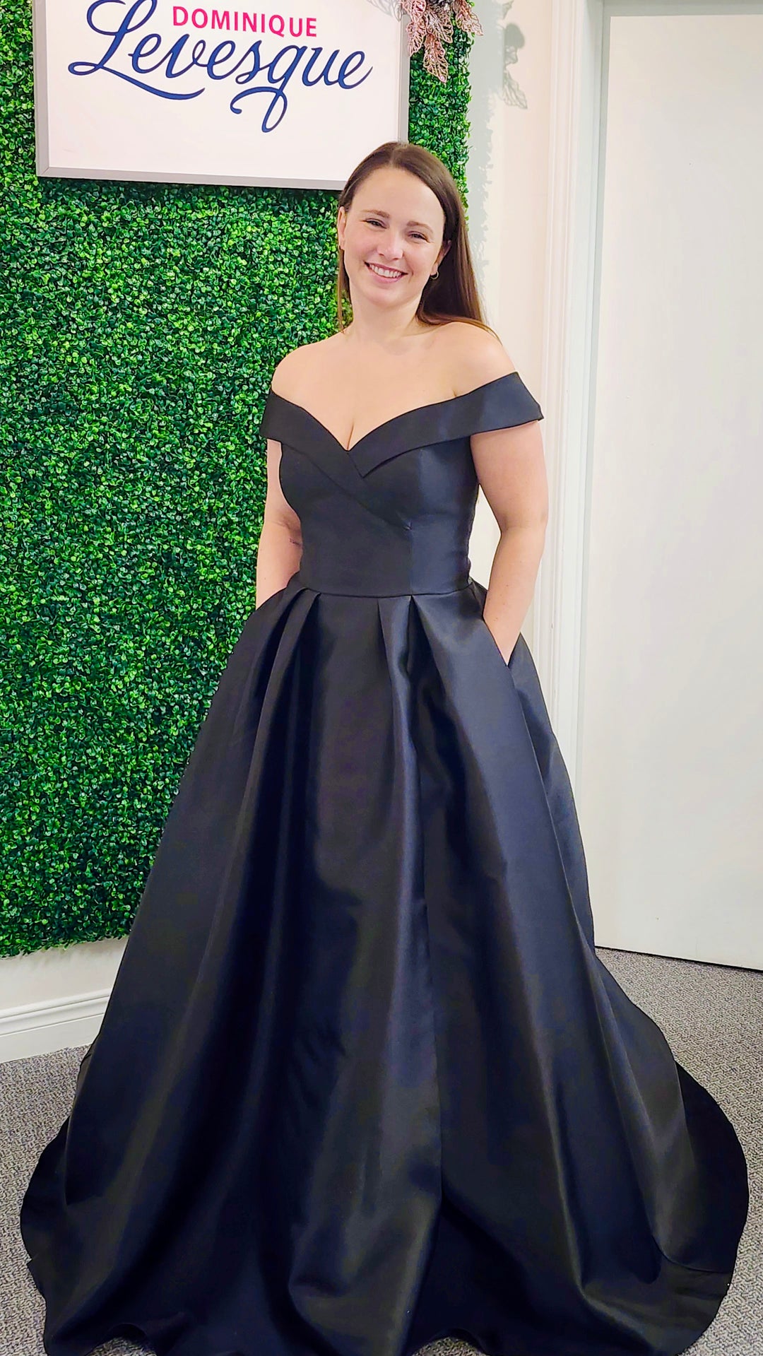 Woman in a navy blue off-shoulder gown standing against a green wall with a sign.