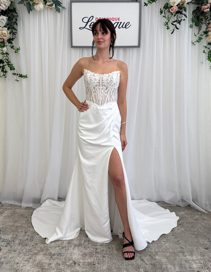 Woman wearing a white strapless wedding dress with a slit in front, standing against a white curtain backdrop.