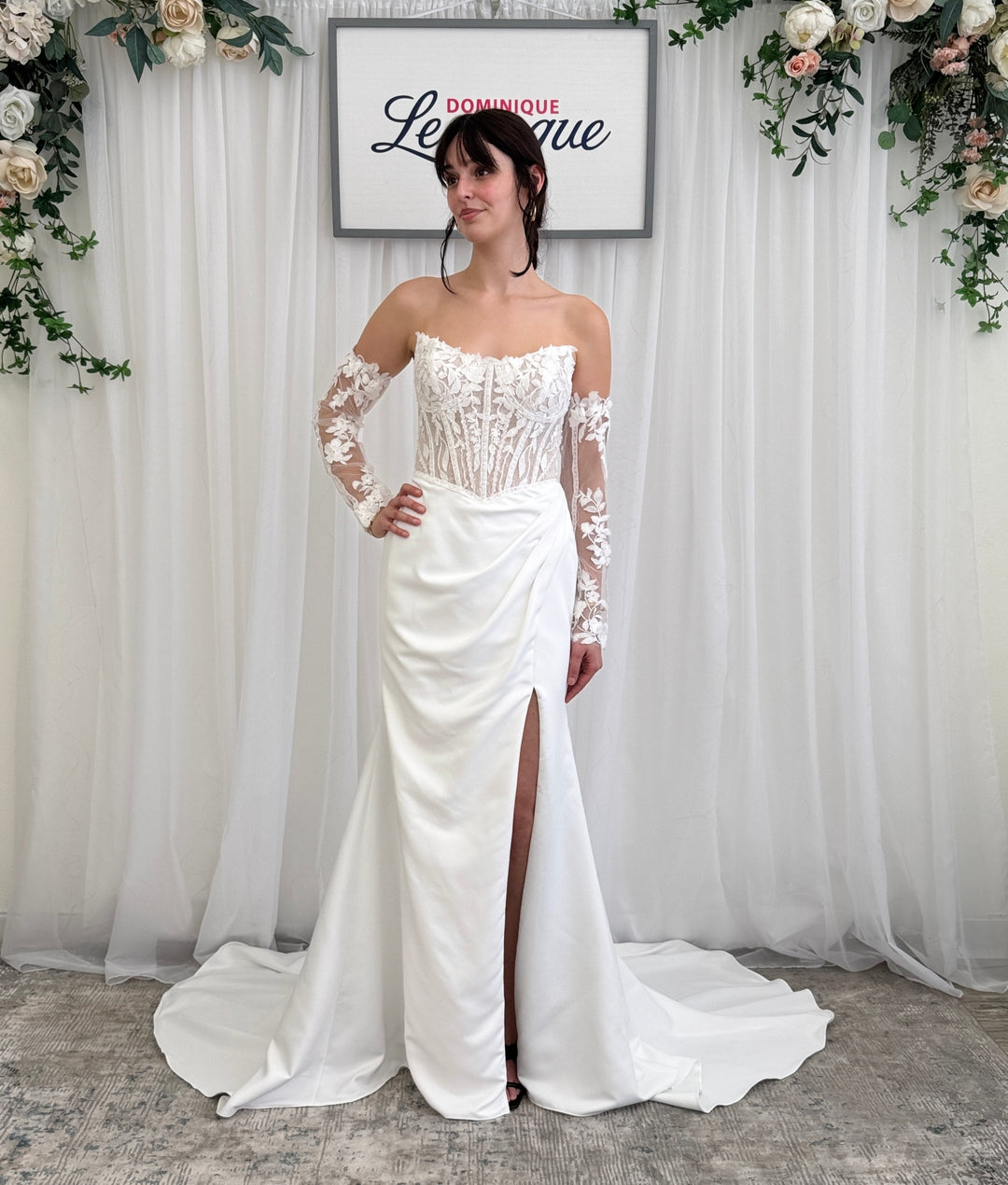 Woman in a white wedding dress with lace sleeves standing in front of a floral backdrop.