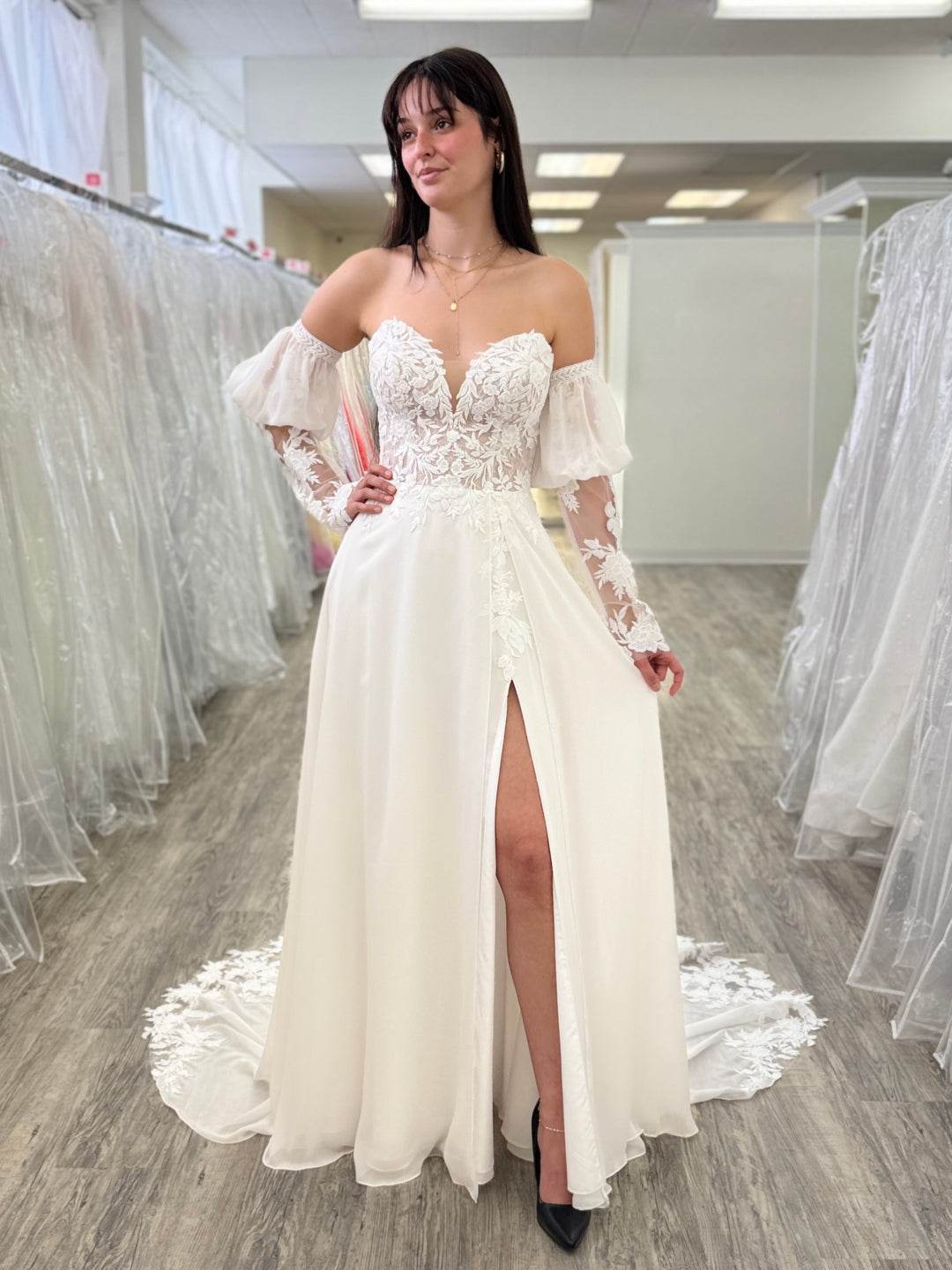 Woman wearing a white wedding dress with lace detailing and a high slit in a fitting room.