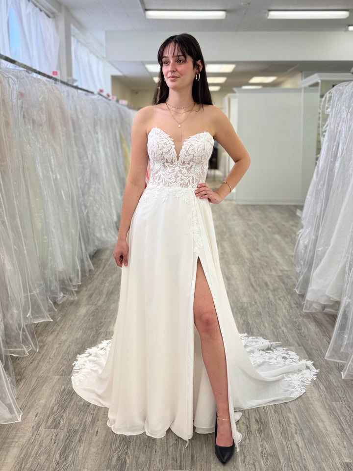 Woman wearing a white wedding dress with lace detailing in a store setting.