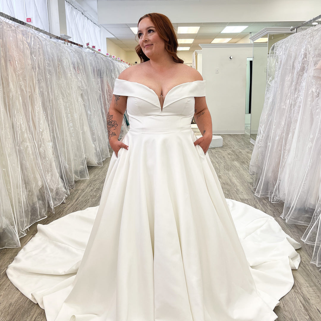 Woman wearing a white off-shoulder wedding dress in a bridal shop.