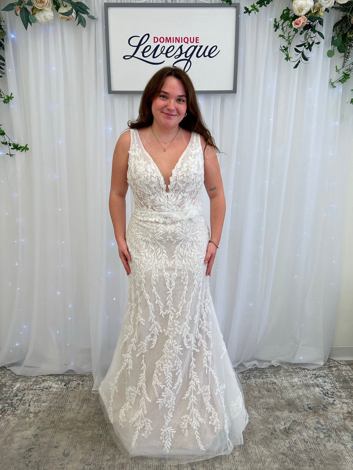 Woman in a white lace dress standing in front of a floral arrangement and framed picture.
