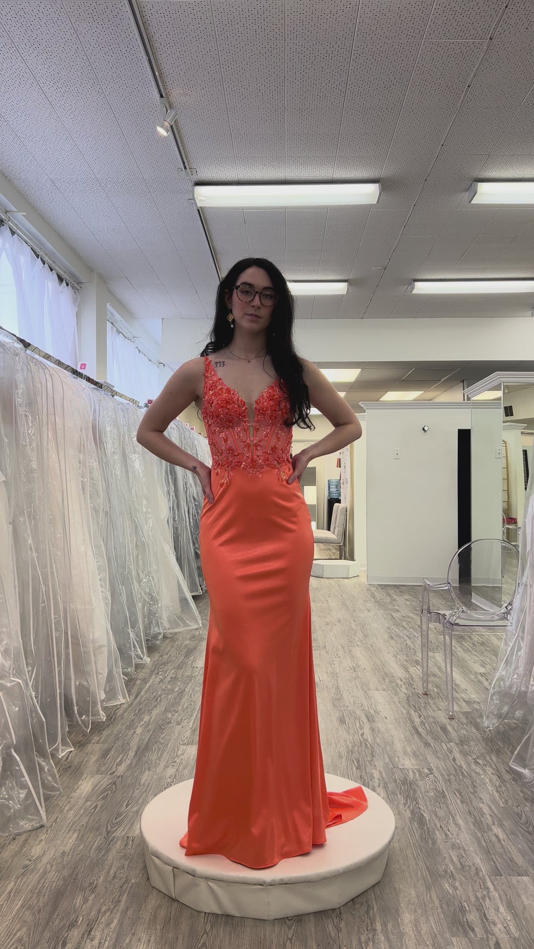 Woman wearing an orange evening gown with lace details in a fitting room.