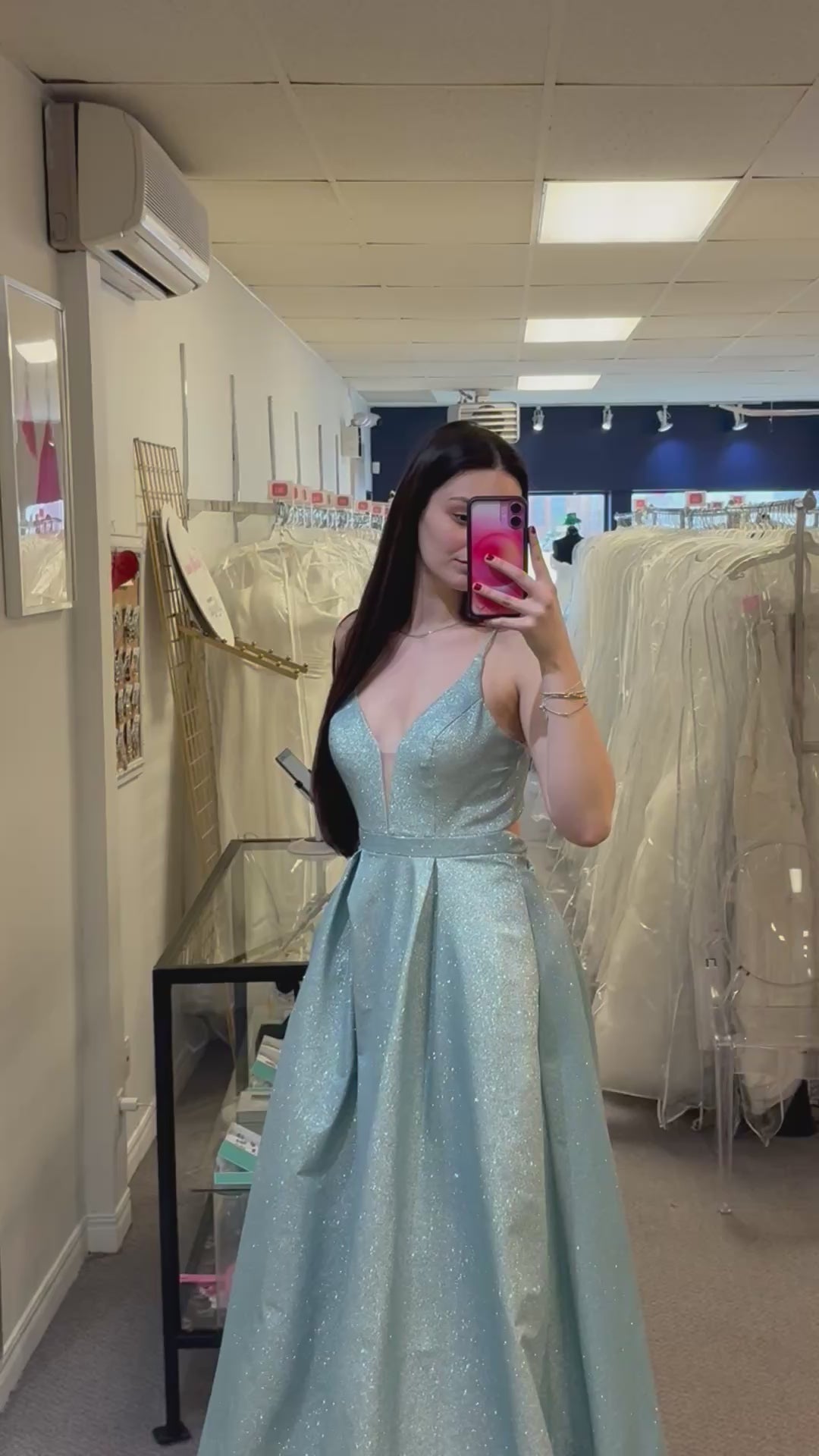 Woman in a light blue evening gown taking a mirror selfie in a fitting room.