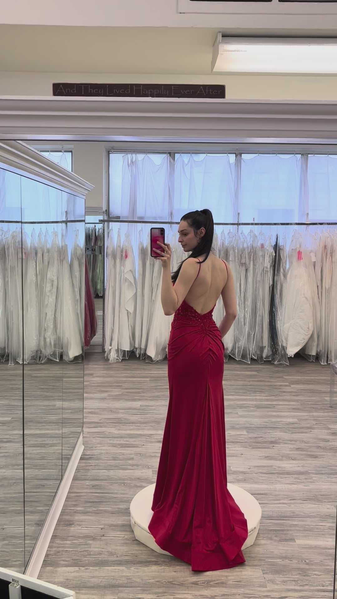 Woman in a red evening gown taking a mirror selfie in a fitting room.
