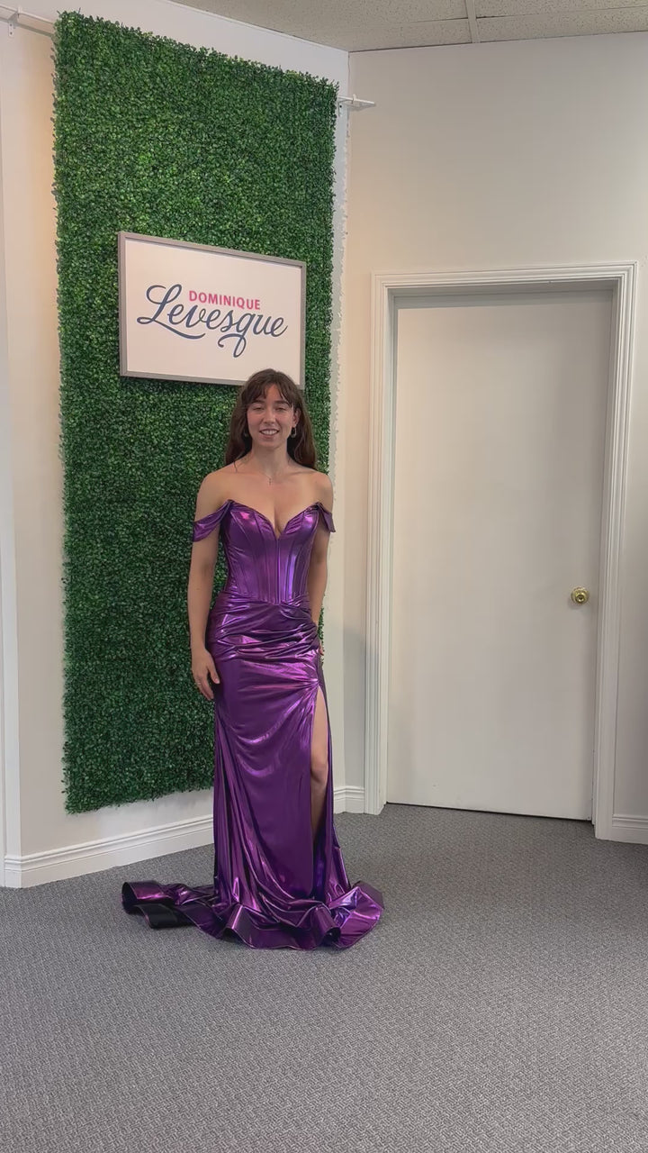 Woman in a purple off-shoulder gown with a high slit standing against a green leafy wall.