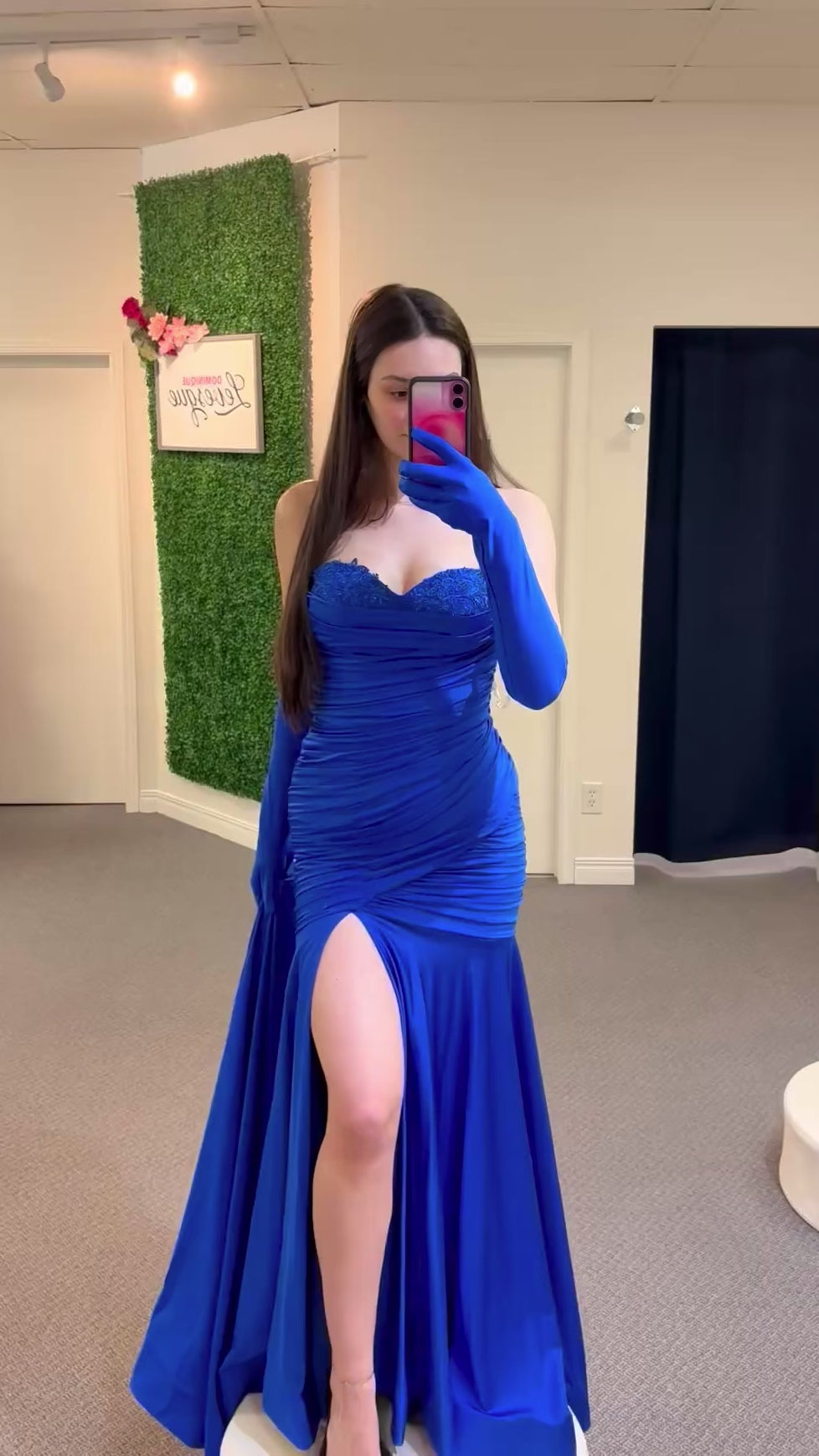 Woman wearing a blue evening gown with a high slit, standing in a fitting room.