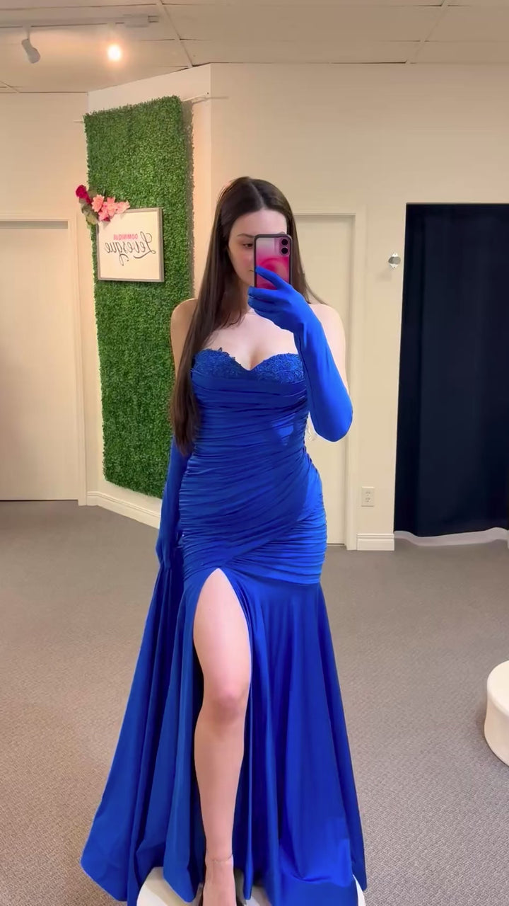 Woman wearing a blue evening gown with a high slit, standing in a fitting room.