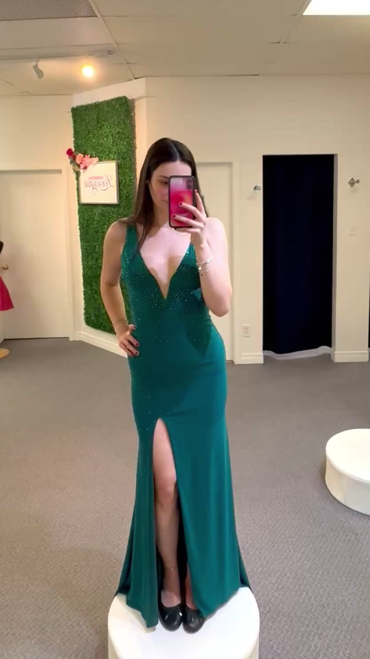 Woman taking a mirror selfie wearing a teal evening gown with a high slit in a fitting room.